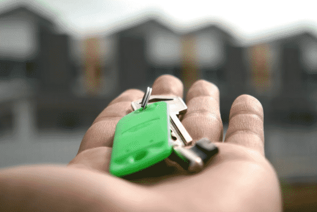 imagem ampliada de pessoa segurando uma chave com chaveiro verde na mão com casas desfocadas no fundo